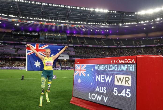 A female athlete, who is wearing running blades, is posing with the Australian flag next to a red electronic board that says "New WR"